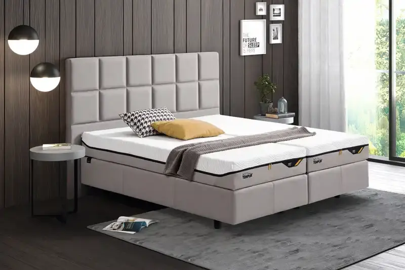 Graues Boxspringbett mit hohem gepolstertem Kopfteil und heller Bettw&auml;sche
